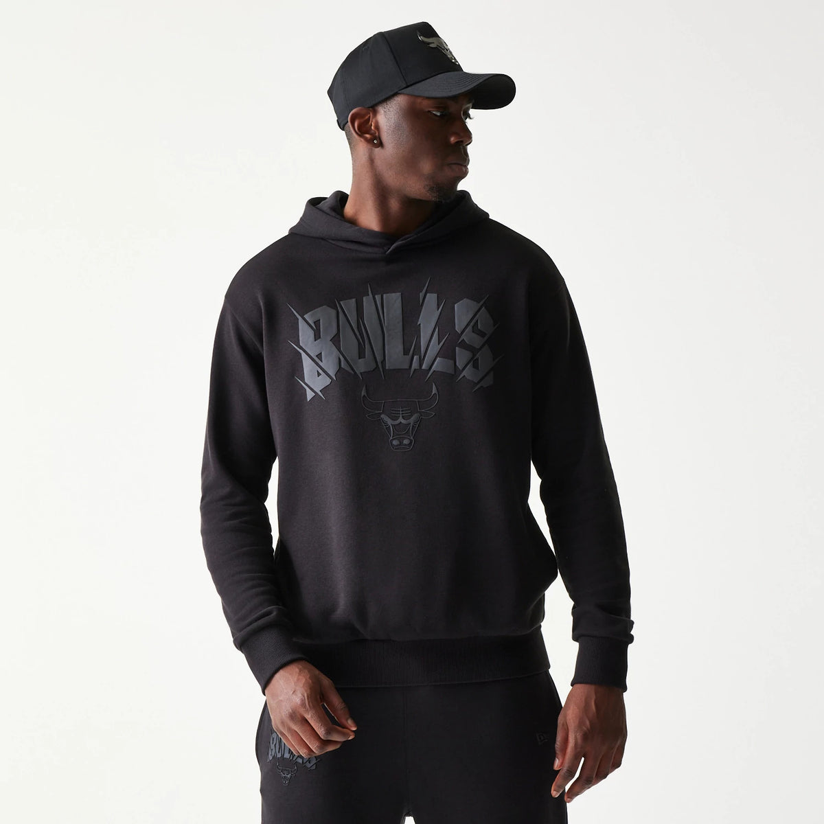 New Era Chicago Bulls NBA Black Oversized Pullover Hoodie, front view showing raised Bulls lettering and the Bulls logo in dark grey across the chest on the black fabric, styled casually with matching tracksuit bottoms.