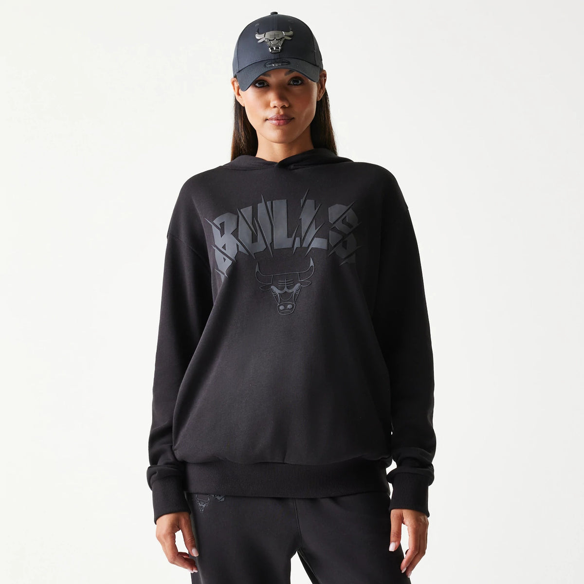 New Era Chicago Bulls NBA Black Oversized Pullover Hoodie, front view on-model showing Bulls lettering and logo on the chest, paired with matching joggers, and styled with a dark cap featuring an embroidered team logo.