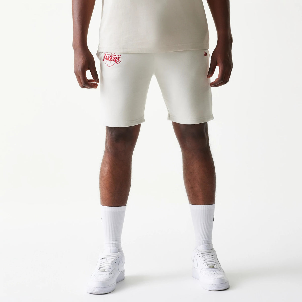 New Era LA Lakers NBA League Essential Cream Shorts, front view showing red embroidered Lakers logo on the left thigh, styled on-model with a neutral top and white trainers, highlighting the clean lines and knee-length fit.