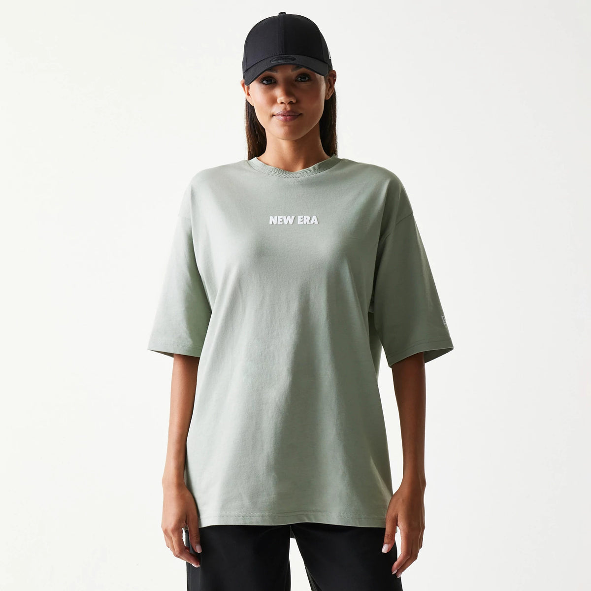 New Era Graphic Pastel Green Oversized T-Shirt, front view on-model showing a relaxed fit with a white printed New Era logo centred on the chest and styled casually with a black cap and dark trousers.