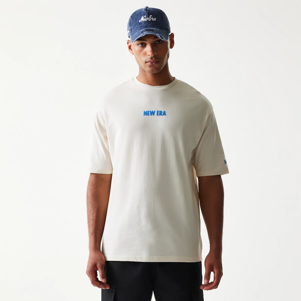 New Era Graphic Light Beige Oversized T-Shirt, front view on-model showing a blue printed New Era logo centred on the chest, short sleeves, and sleeve embroidery, styled with a blue cap.