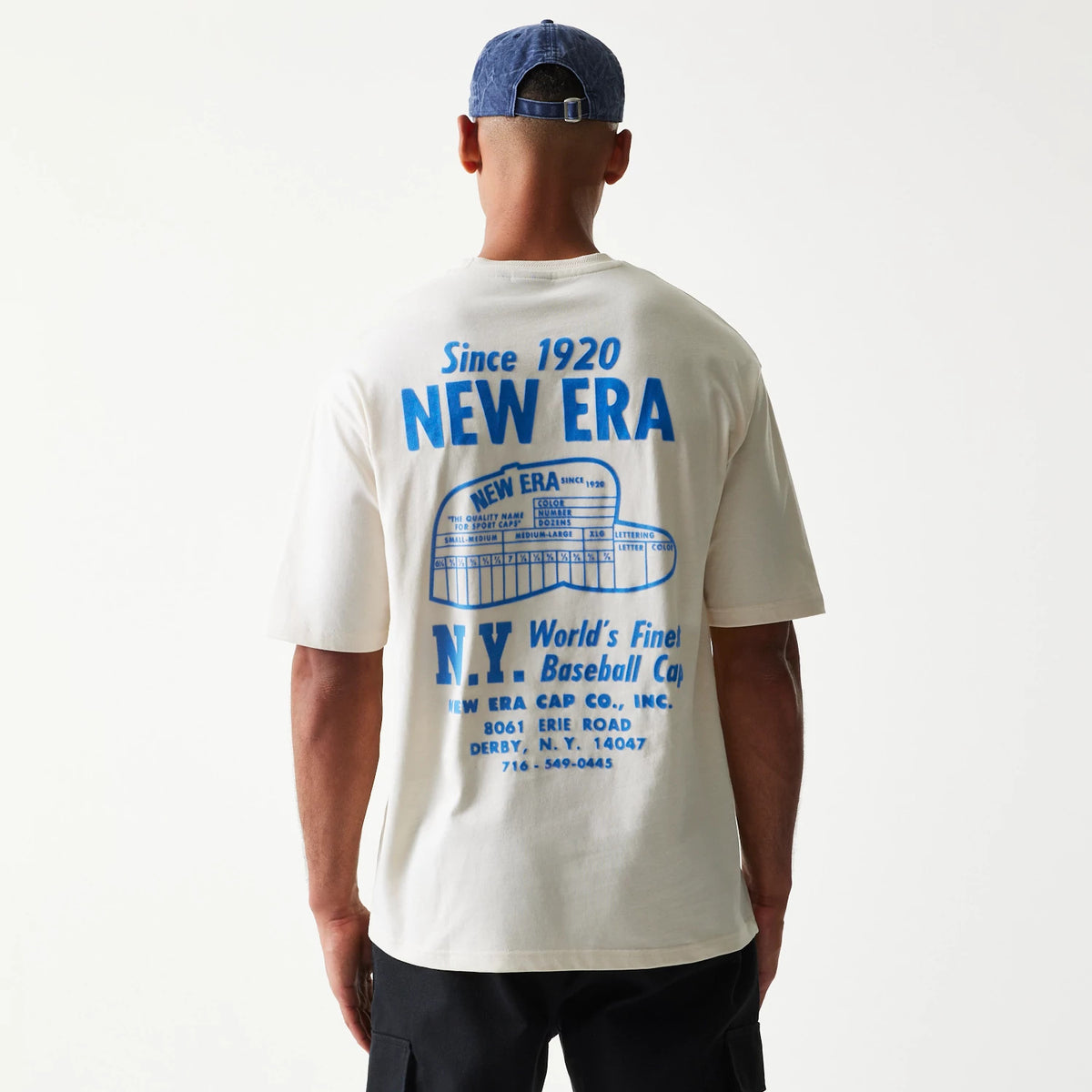 New Era Graphic Light Beige Oversized T-Shirt, back view on a model with a blue cap, displaying a large blue printed graphic featuring Since 1920 New Era lettering and a stylised baseball cap image with additional text across the back.