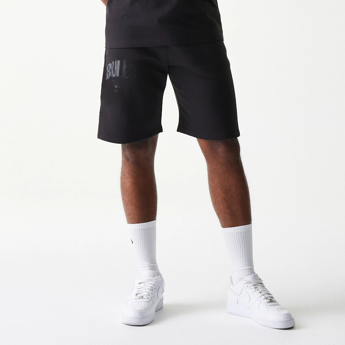New Era Chicago Bulls NBA Black Shorts, front view on-model showing the black shorts with an embroidered team logo and text on the left leg, styled with white socks and trainers for a casual look.