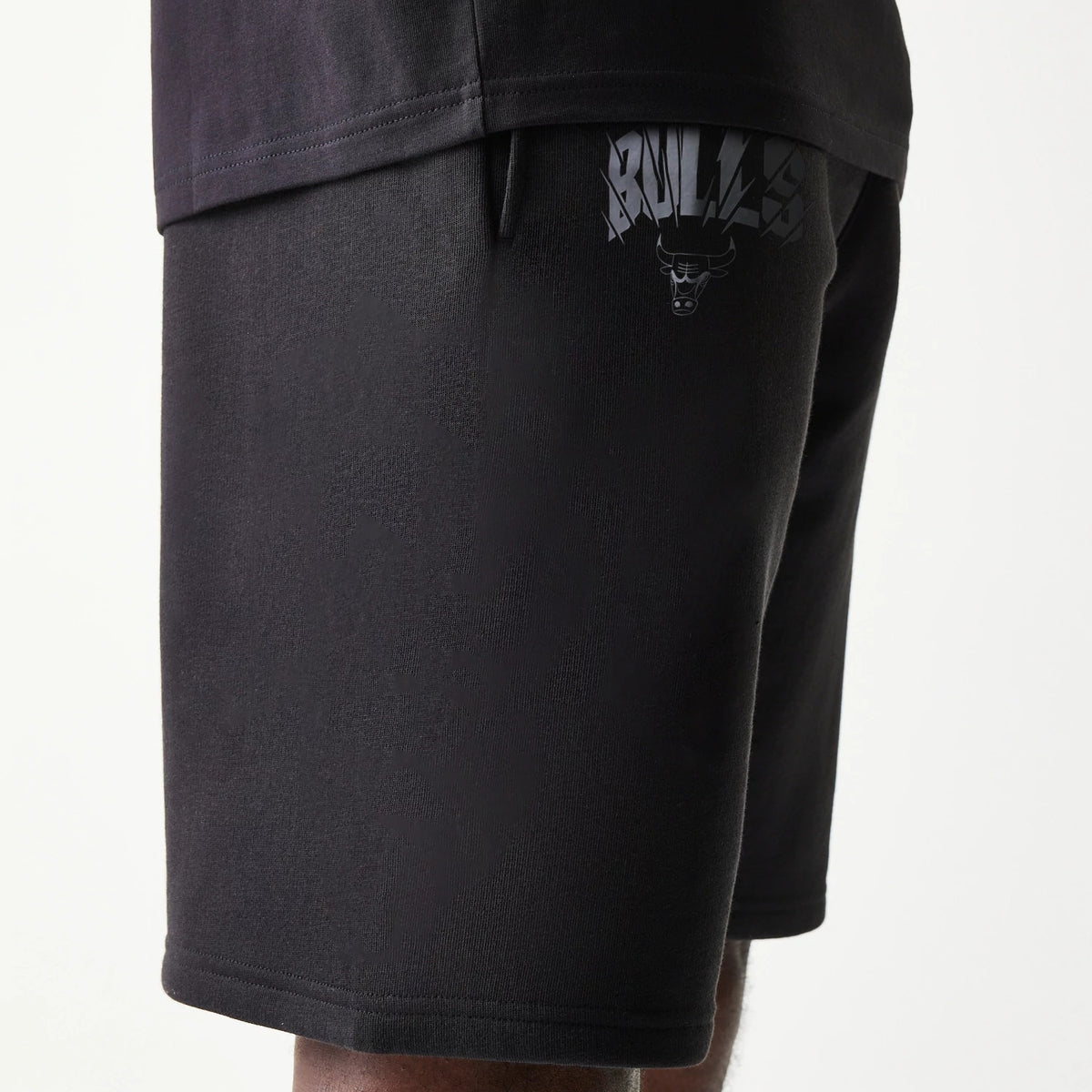 New Era Chicago Bulls NBA Black Shorts, side view showing the shorts worn with a black top, featuring an embossed Bulls logo and text on the lower side of the black fabric, creating a and athletic look.