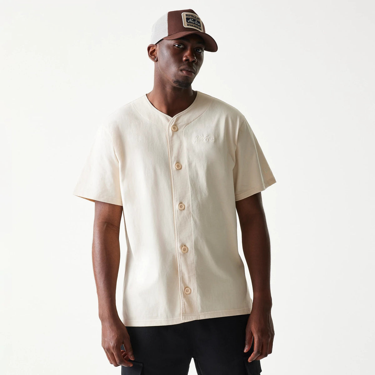 New Era Script Light Beige T-Shirt, front view on-model showing a classic button-down design with an embroidered New Era logo on the left chest, paired with a contrasting brown and white cap.