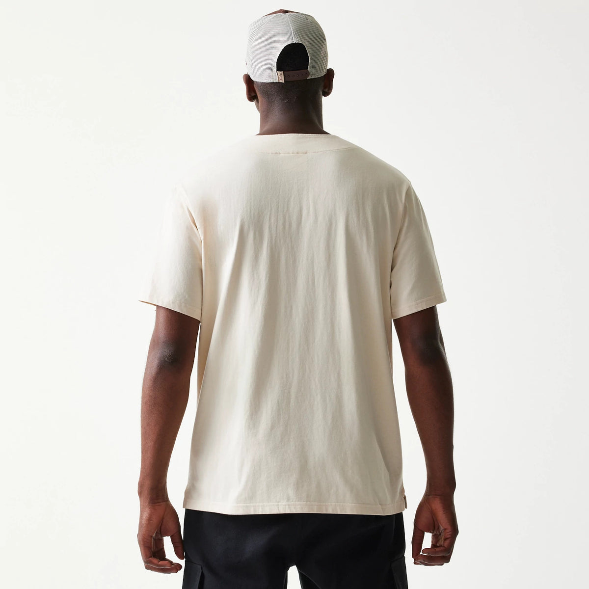 New Era Script Light Beige T-Shirt, back view on a model with a plain light beige surface and short sleeves, showcasing the minimal design and relaxed fit worn with black trousers and a light-coloured cap.