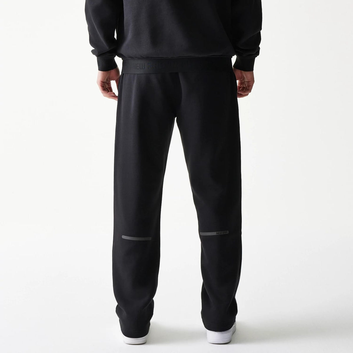 New Era Tech Black on Black Straight Leg Joggers, back view on a model showing the elastic waistband with branding, straight leg fit with grey horizontal accents above the knees, and white trainers.