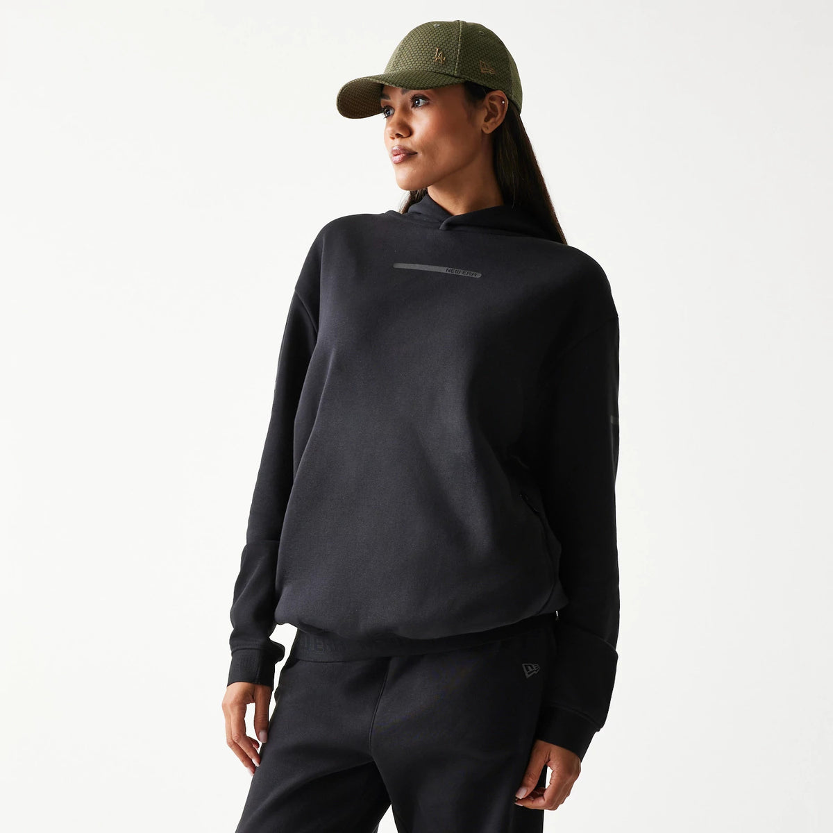 New Era Tech Strip Black Oversized Pullover Hoodie, front view on a model styled with dark trousers and a green cap, featuring a black printed strip across the chest and simple cuffed sleeves.