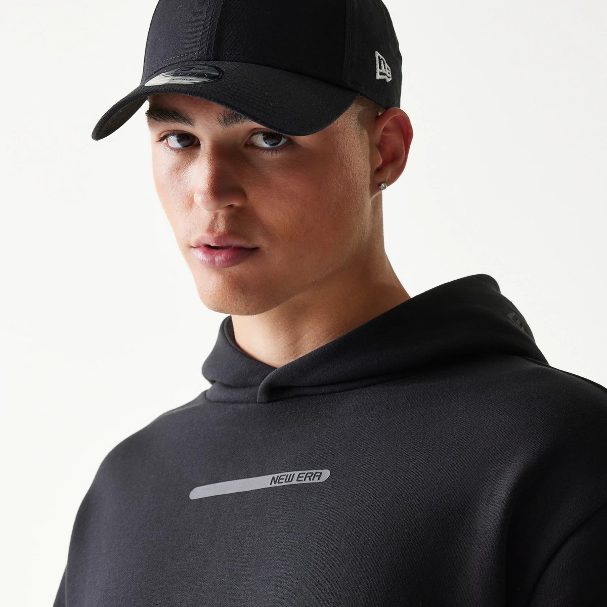 New Era Tech Strip Black Oversized Pullover Hoodie, front view on-model showing the grey printed New Era text with a strip on the chest below the hood, paired with a black cap featuring an embroidered side logo.