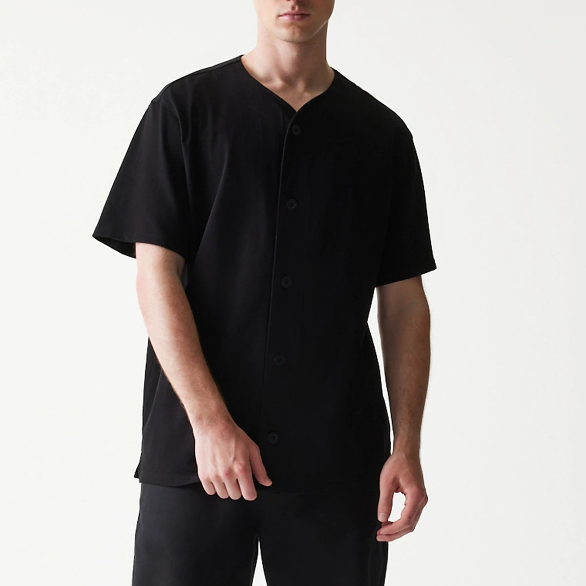 New Era Open Heritage Script Black T-Shirt, front view on-model showing the plain black design with a button-down front, short sleeves, and a round neckline, styled simply with matching black trousers.