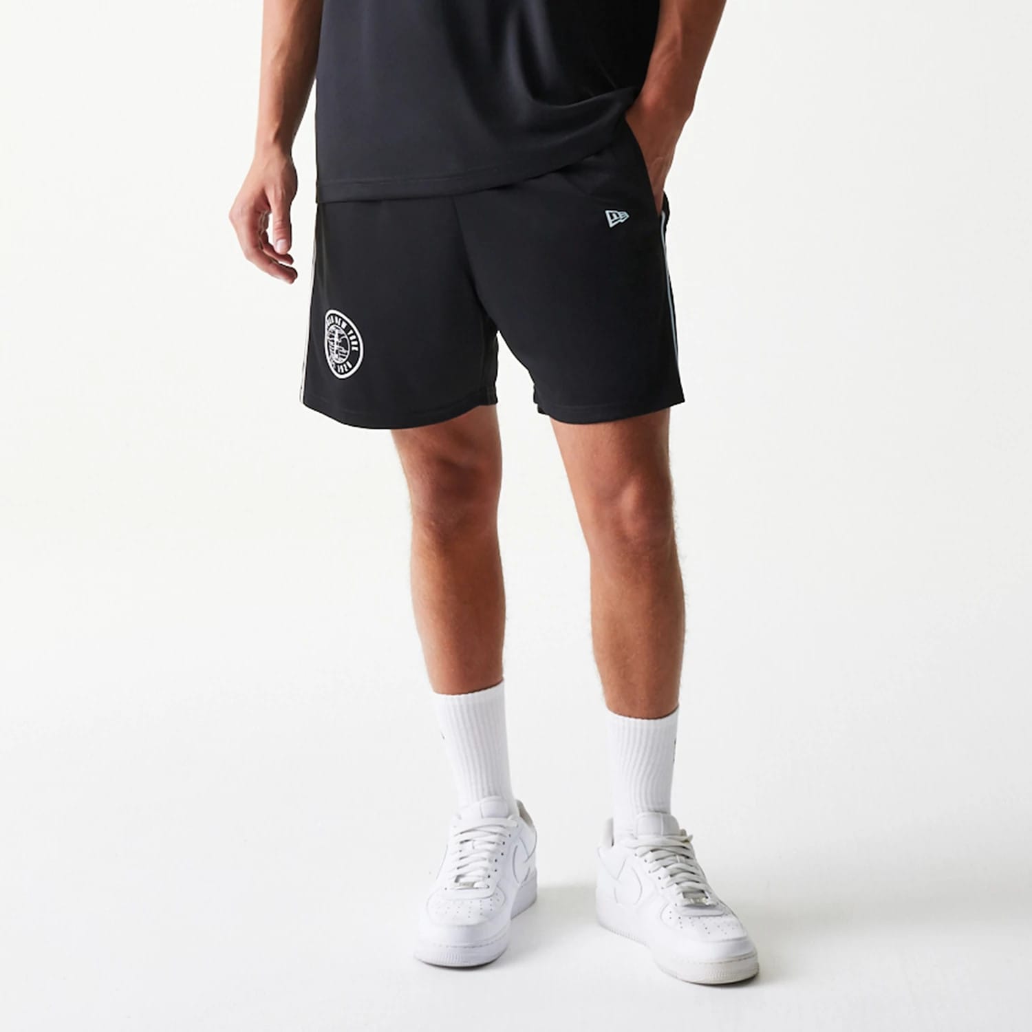 New Era Football Black Shorts, front view on a model showing the embroidered New Era logo on the left leg and a circular team graphic on the right leg, styled with a black top and white trainers.