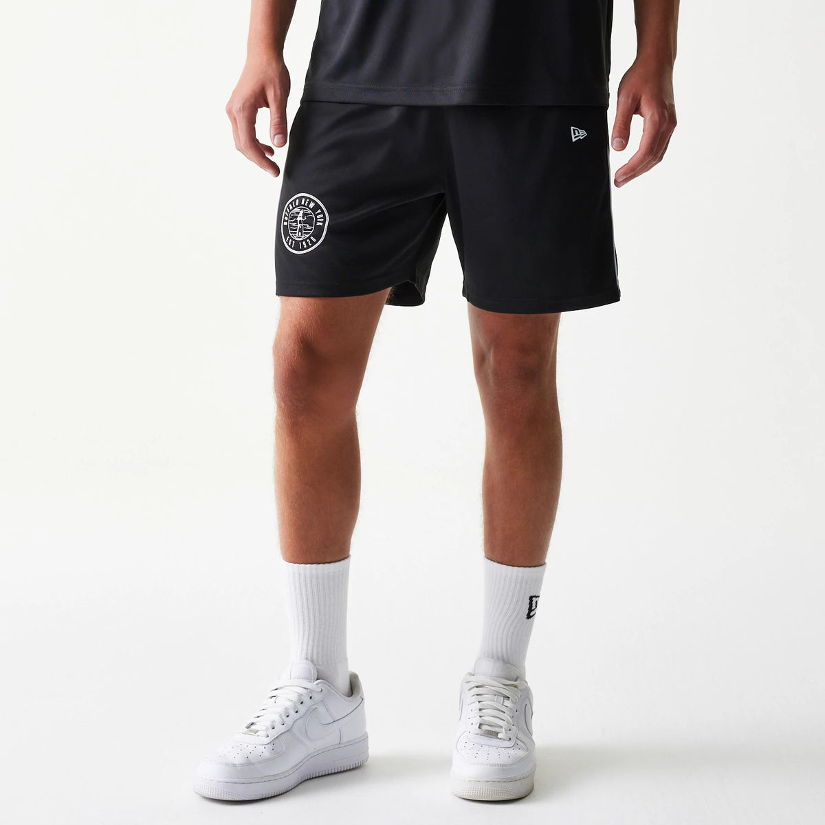 New Era Football Black Shorts, front view on a model showing a round monochrome team graphic on the left leg near the hem and a small white embroidered New Era logo positioned higher on the opposite side.