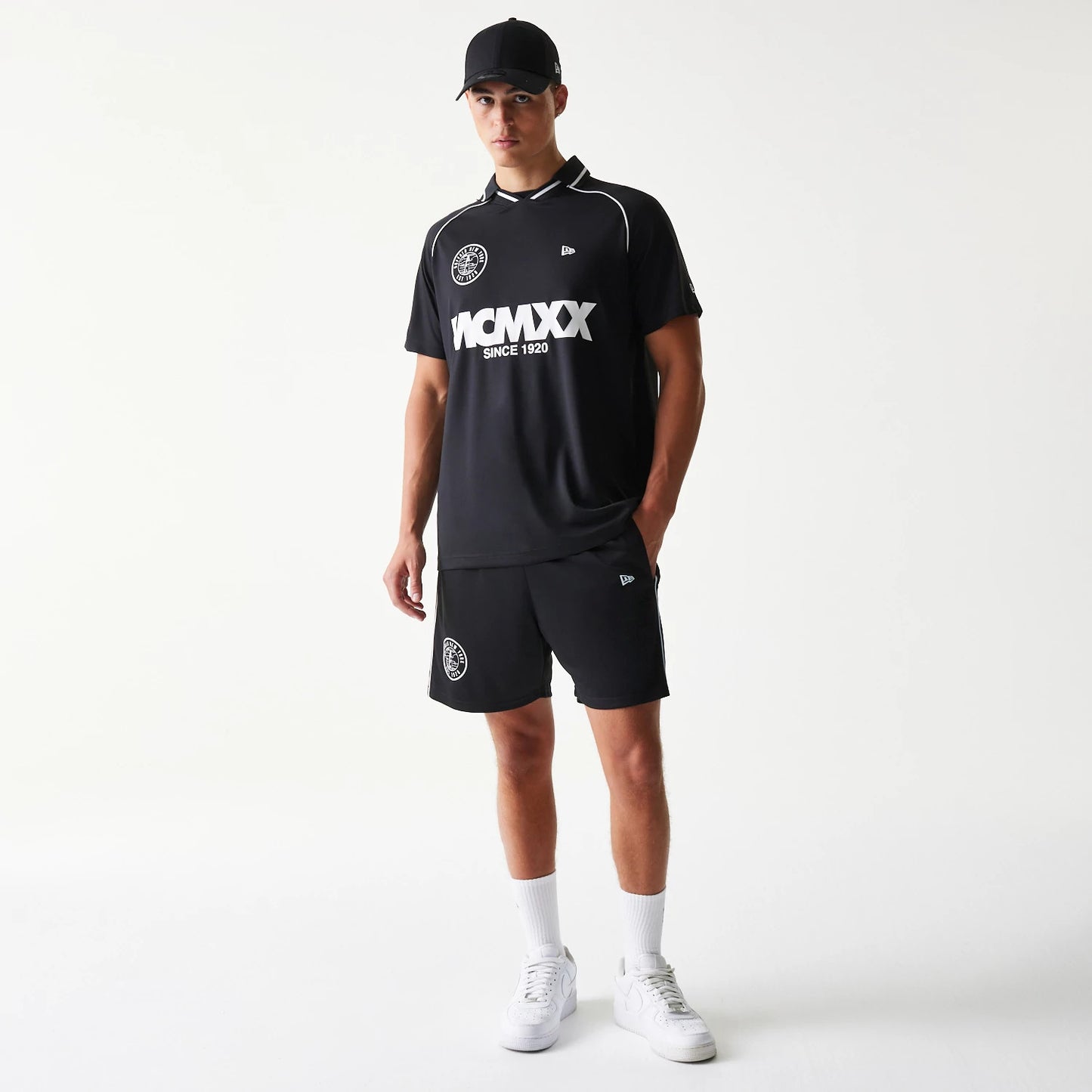 New Era Football Black Shorts, front view on a model with a matching black top and cap, showing a white circular graphic on the left leg and an embroidered New Era logo on the right, above the hem.