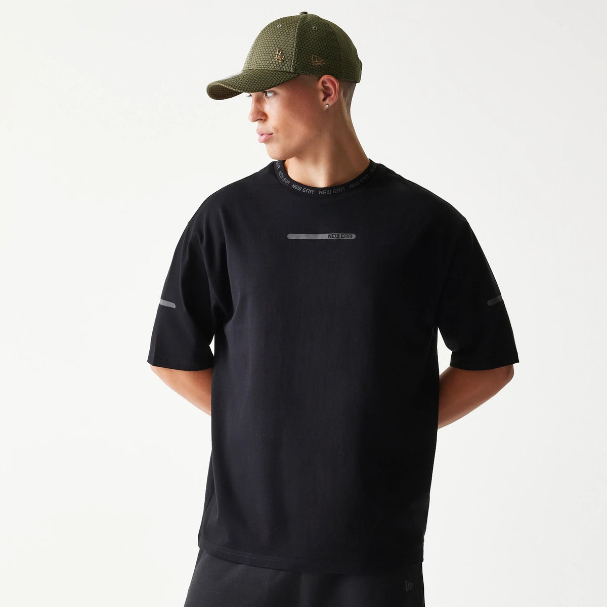 New Era Tech Black Oversized T-Shirt, front view on-model showing the New Era logo printed across the chest and neckline, with short sleeves featuring reflective stripes on each arm and relaxed fit styling.