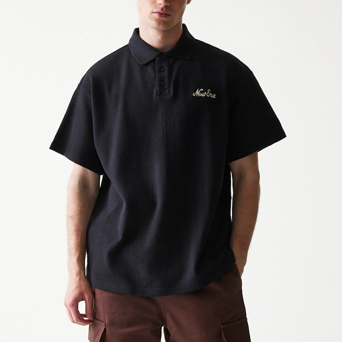 New Era Graphic Black Polo T-Shirt, front view showing the classic polo style with a button placket, short sleeves, and an embroidered New Era logo on the left chest, styled on a model with casual brown trousers.