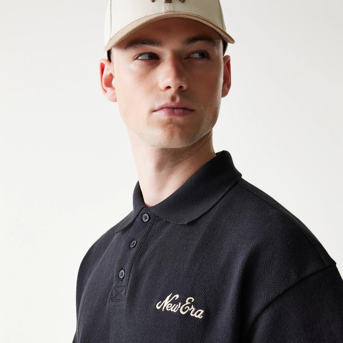 New Era Graphic Black Polo T-Shirt, three-quarter view showing the black polo with a embroidered New Era logo on the left chest, worn by a model also styled with a neutral-toned cap.