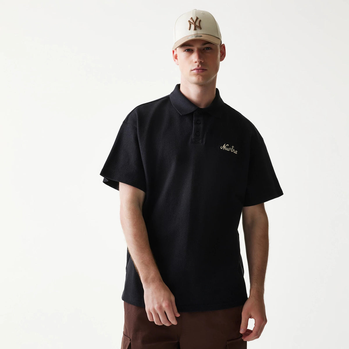 New Era Graphic Black Polo T-Shirt, front view on-model showing the classic polo collar with three buttons and an embroidered New Era logo on the left chest, styled with brown trousers and a light-coloured cap.