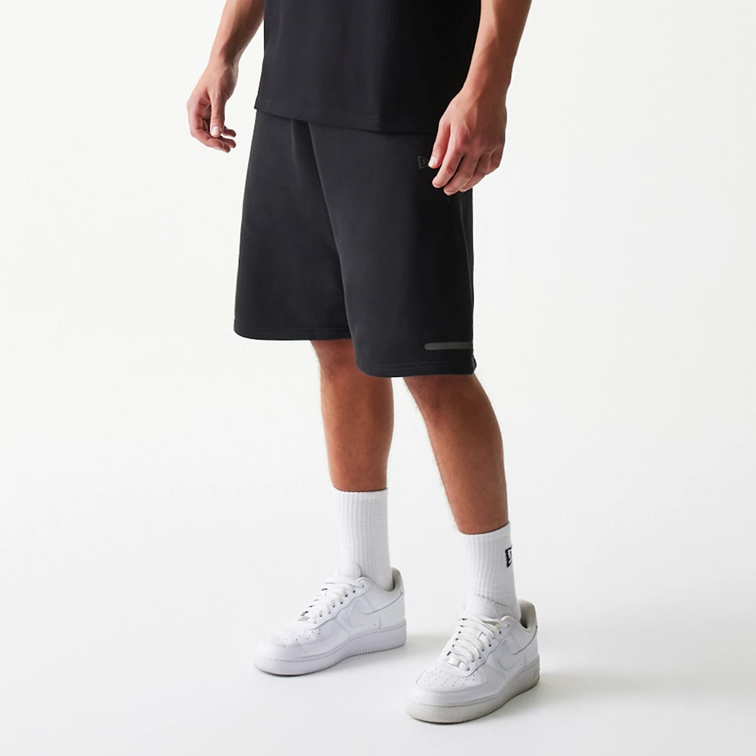 New Era Tech Black Shorts, front view showing the relaxed fit with knee-length cut, a small embroidered logo on the left leg, and a reflective strip near the hem, styled casually with white trainers and socks.
