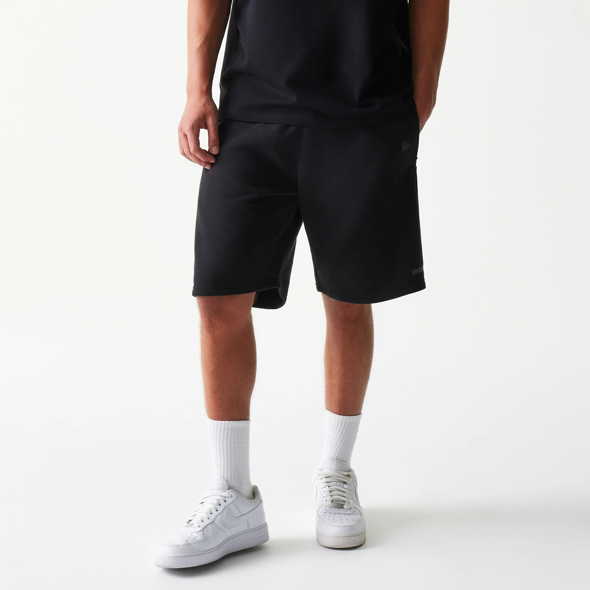 New Era Tech Black Shorts, front view on-model showing knee-length loose fit shorts with an embroidered New Era logo on the lower left leg, styled with a matching black top, white socks, and white trainers.