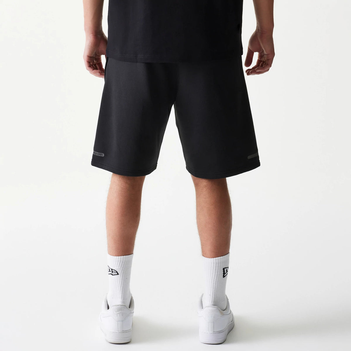 New Era Tech Black Shorts, back view showing the simple black design with a debossed New Era logo on the lower left leg, styled with white socks featuring the New Era logo.