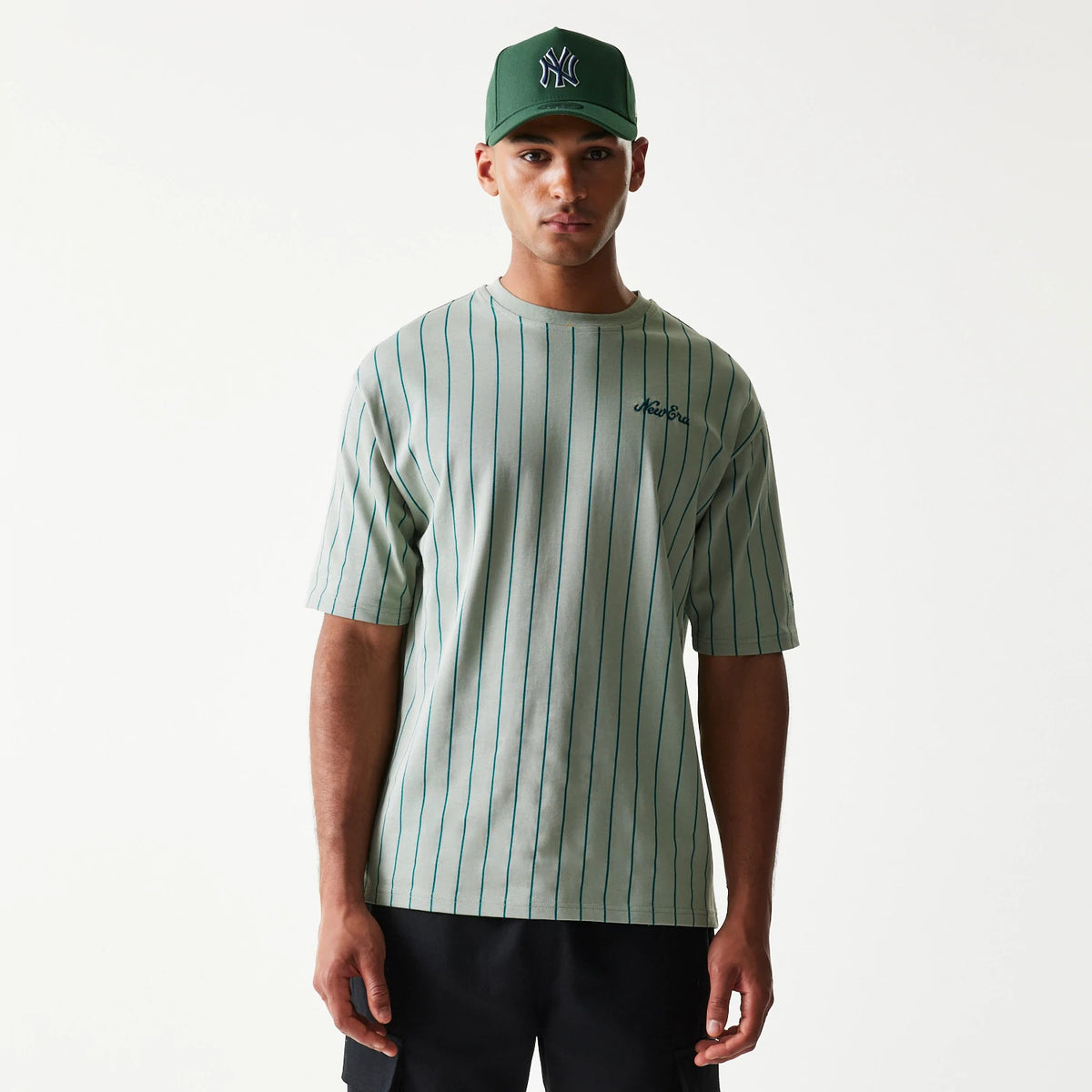 New Era Pinstripe Pastel Green Oversized T-Shirt, front view on a model wearing a matching green cap, displaying thin vertical stripes and an embroidered New Era script logo on the left chest.