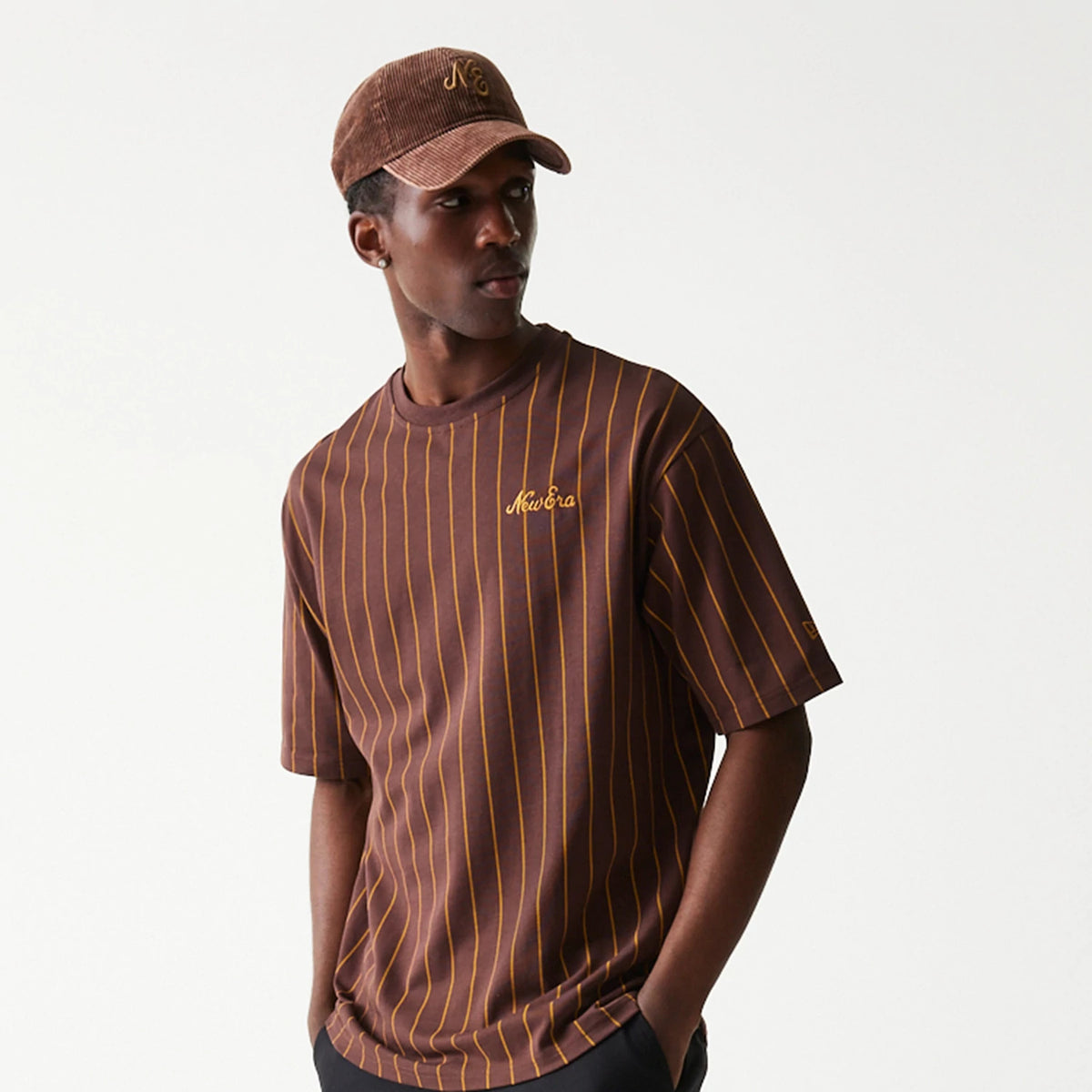 New Era Pinstripe Brown Oversized T-Shirt, front view on-model featuring a brown base with mustard yellow pinstripes, an embroidered New Era logo on the left chest and accompanied by matching brown cap.