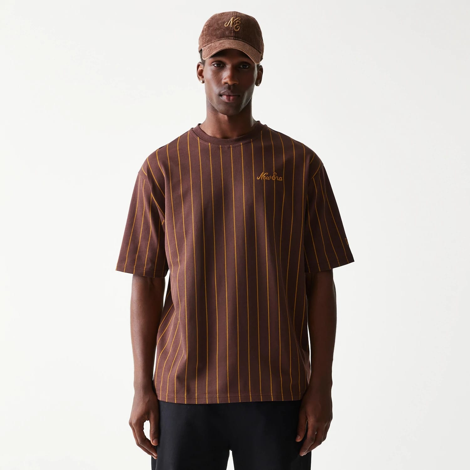 New Era Pinstripe Brown Oversized T-Shirt, front view on-model showing the brown tee with vertical yellow pinstripes, an embroidered New Era logo on the chest, styled with a matching brown cap featuring an embroidered logo.