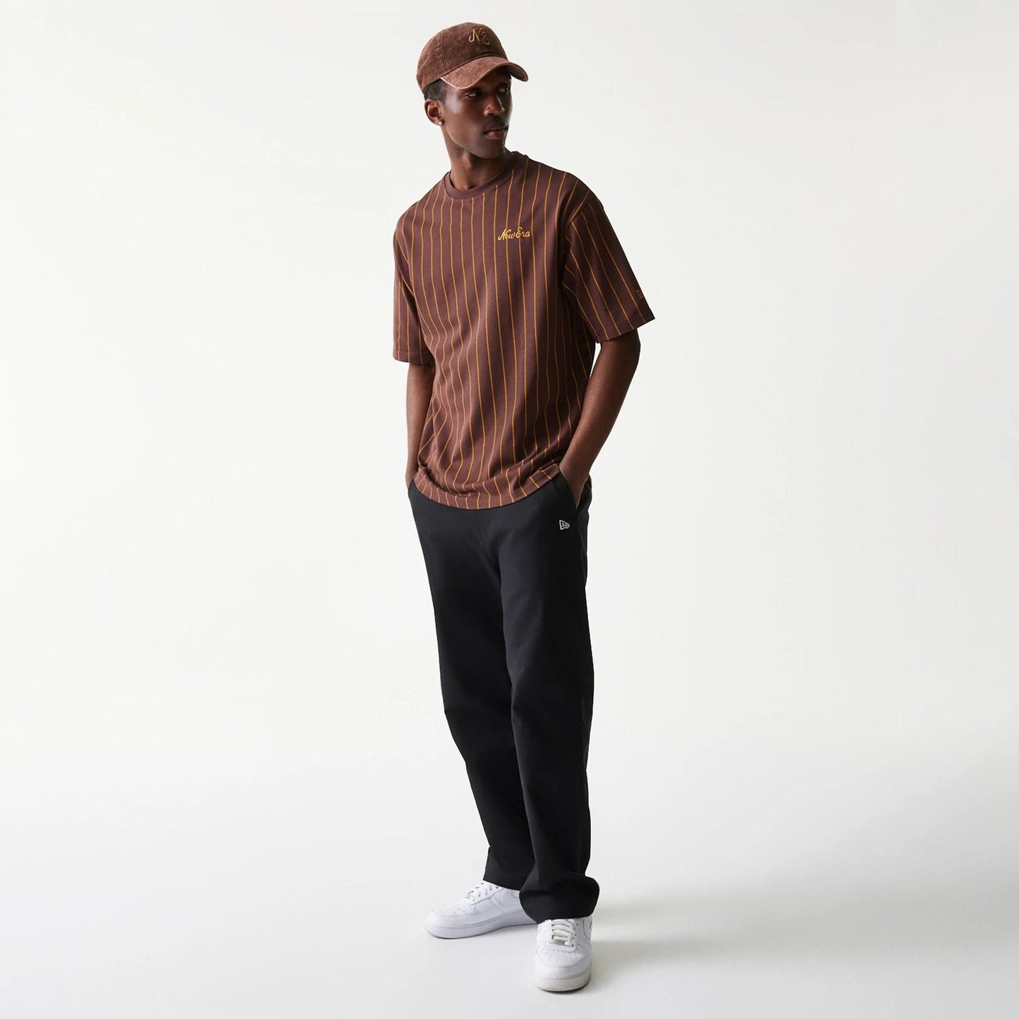 New Era Pinstripe Brown Oversized T-Shirt, front view on-model featuring vertical beige pinstripes, an embroidered yellow logo on the left chest, styled with black trousers and white trainers, completing a casual look.