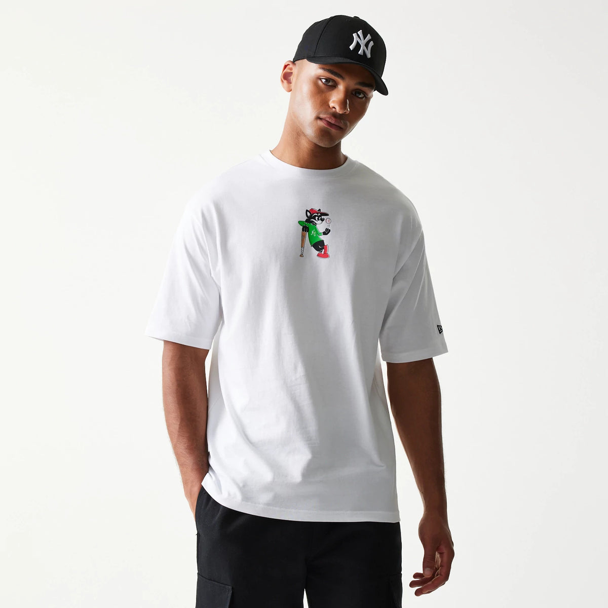 New Era Baseball Cat Character Graphic White Oversized T-Shirt, front view showing a cartoon cat graphic with a bat on the chest and a small embroidered New Era logo on the left sleeve, on-model styled with a black cap.