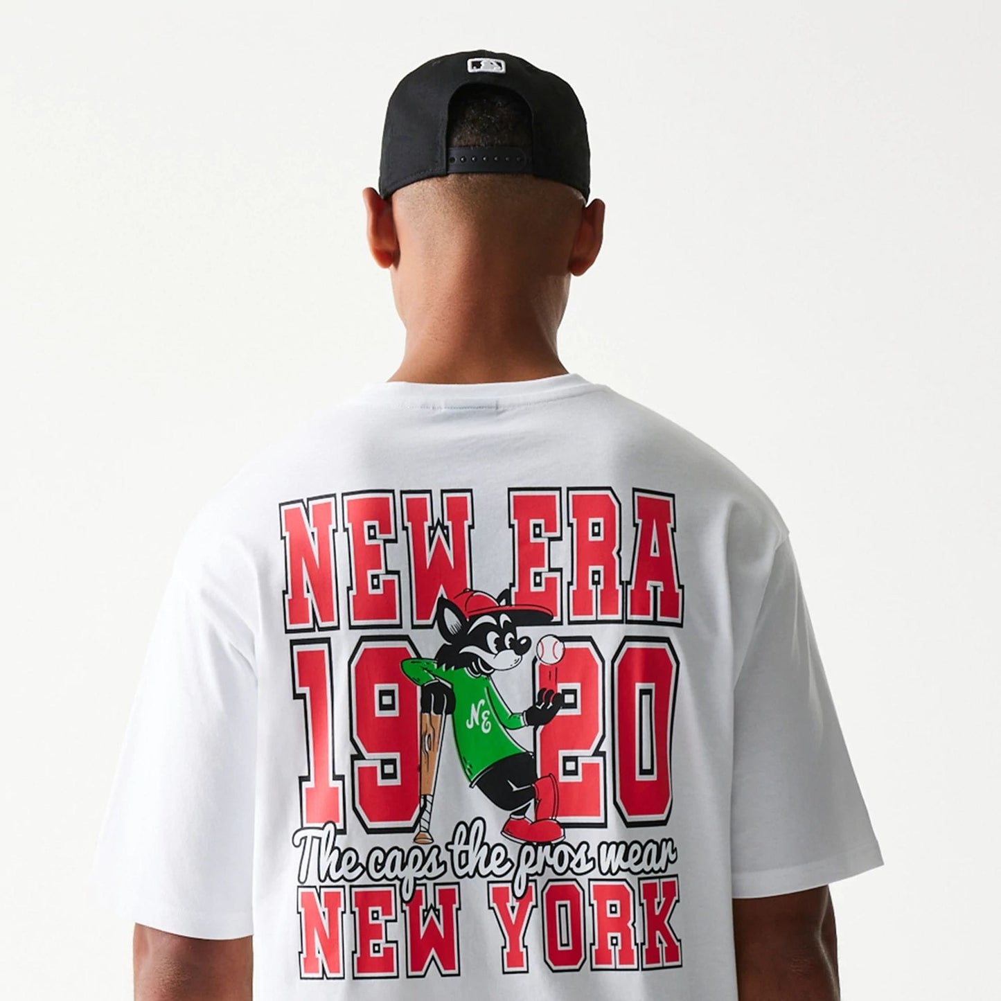 New Era Baseball Cat Character Graphic White Oversized T-Shirt, back view on a model with a black cap, featuring New Era 1920 text and a cartoon cat in a green jersey holding a baseball bat and ball.