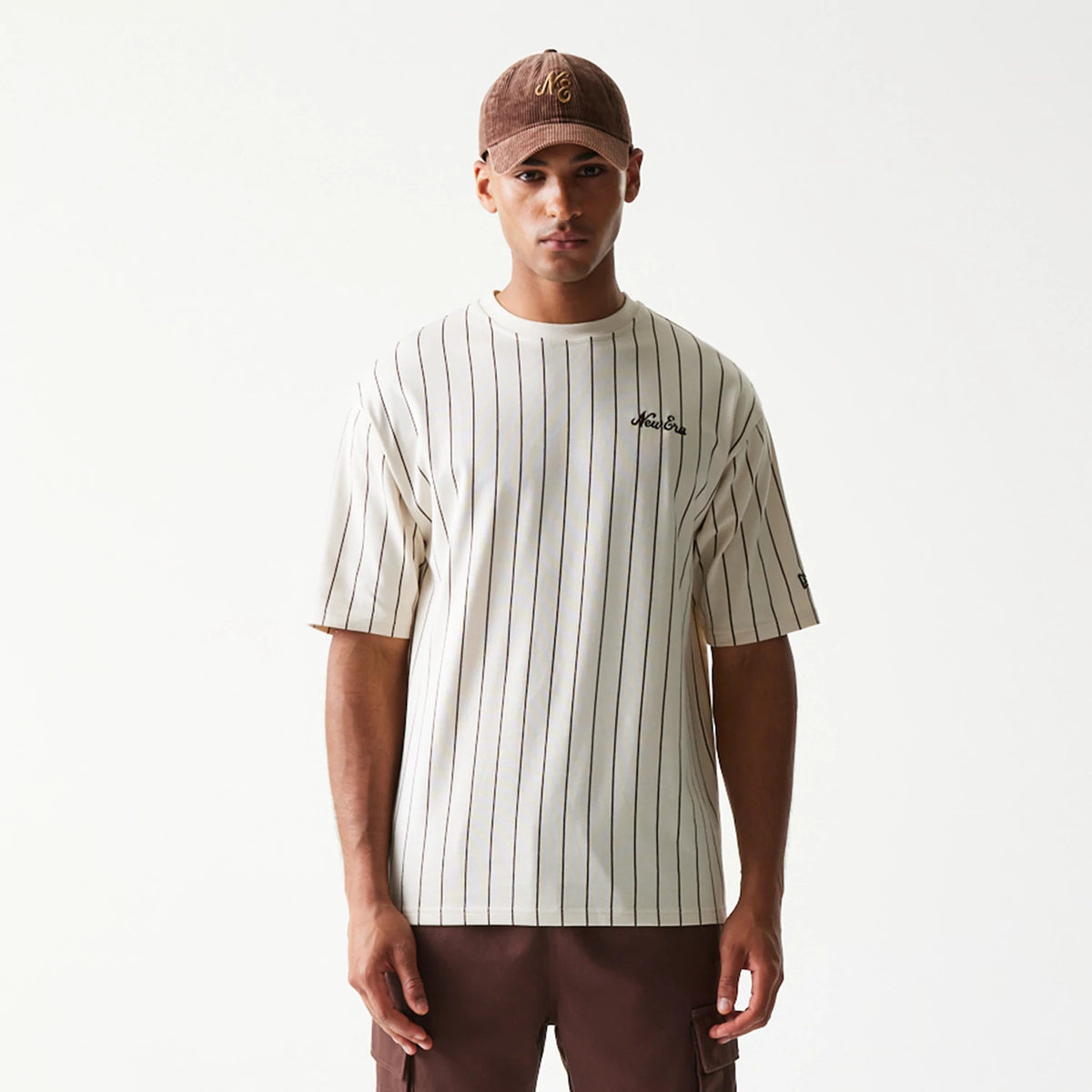 New Era Pinstripe Light Beige Oversized T-Shirt, front view showing vertical pinstripes across the beige fabric, an embroidered New Era logo on the left chest, and a relaxed fit with short sleeves.