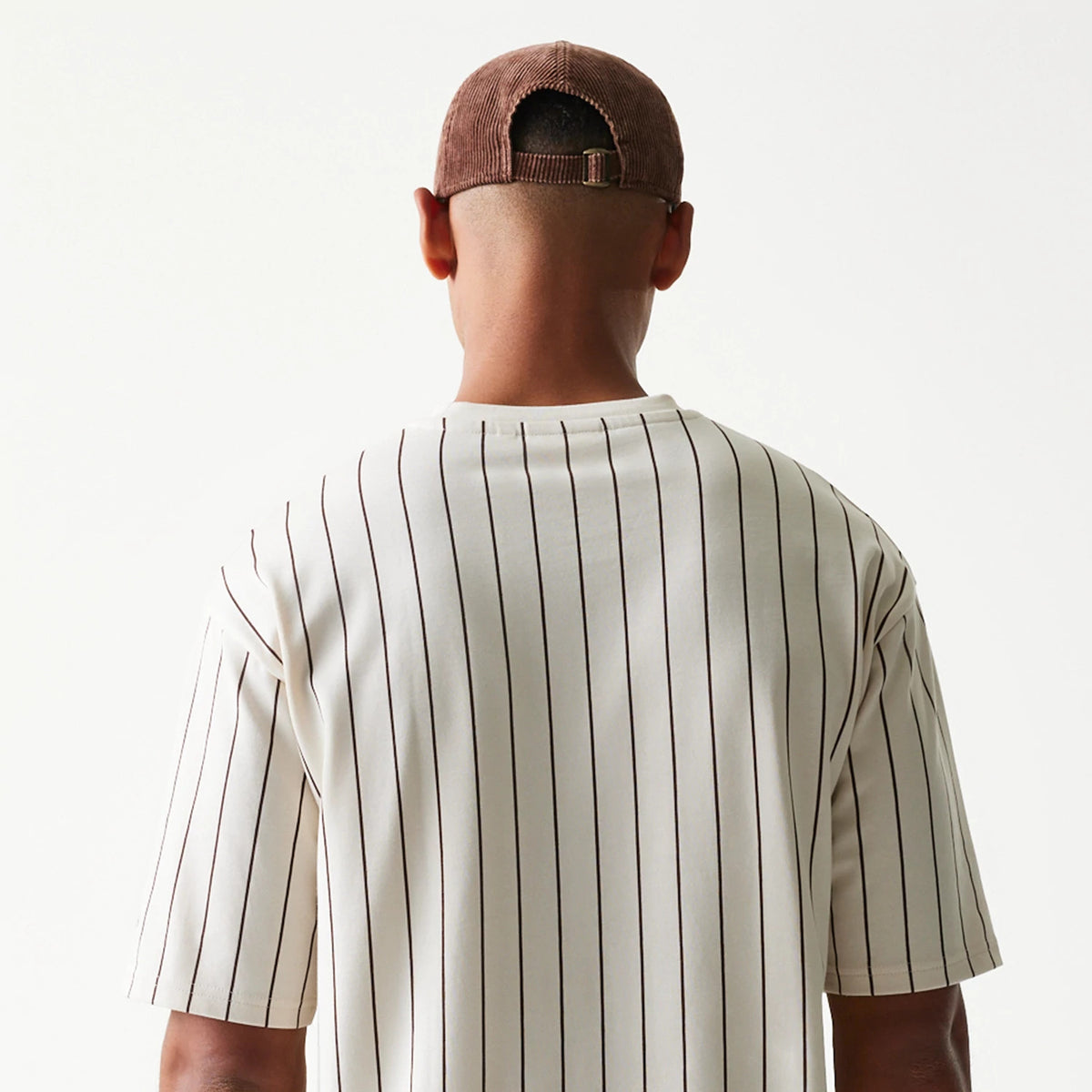 New Era Pinstripe Light Beige Oversized T-Shirt, back view on a model paired with a brown cap, featuring thin vertical pinstripes running across the light beige fabric and short sleeves extending to the elbows.