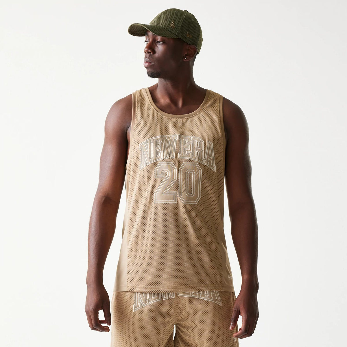 New Era Mesh Light Beige Tank Top, front view on a model showing white embroidered NEW ERA 20 lettering across the chest, with the sleeveless design and a light beige mesh fabric for a casual sporty look.