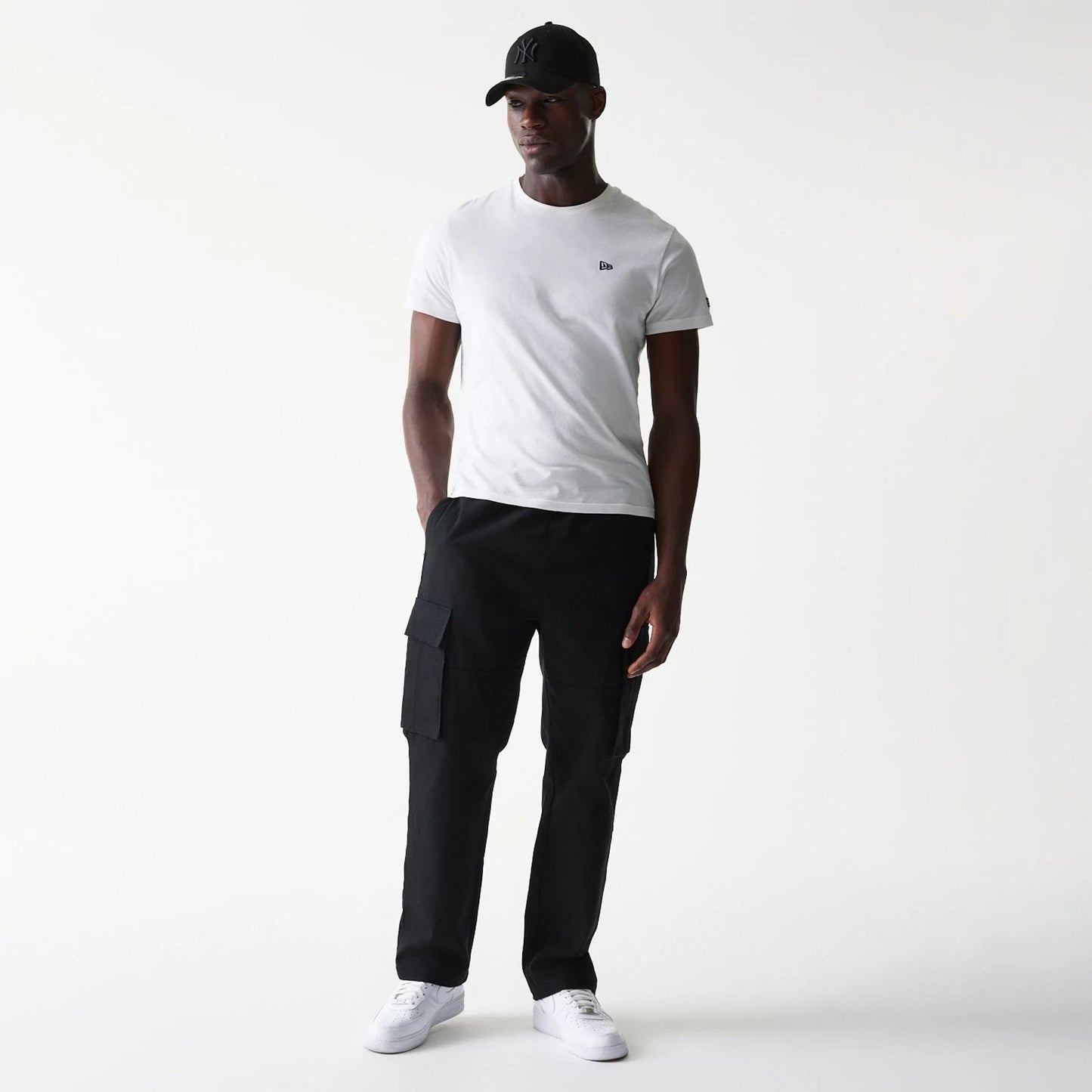 New Era Black Straight Leg Cargo Joggers, front view on a model wearing a white T-shirt and black cap, showing the joggers' side cargo pockets and relaxed fit with hands in pockets.