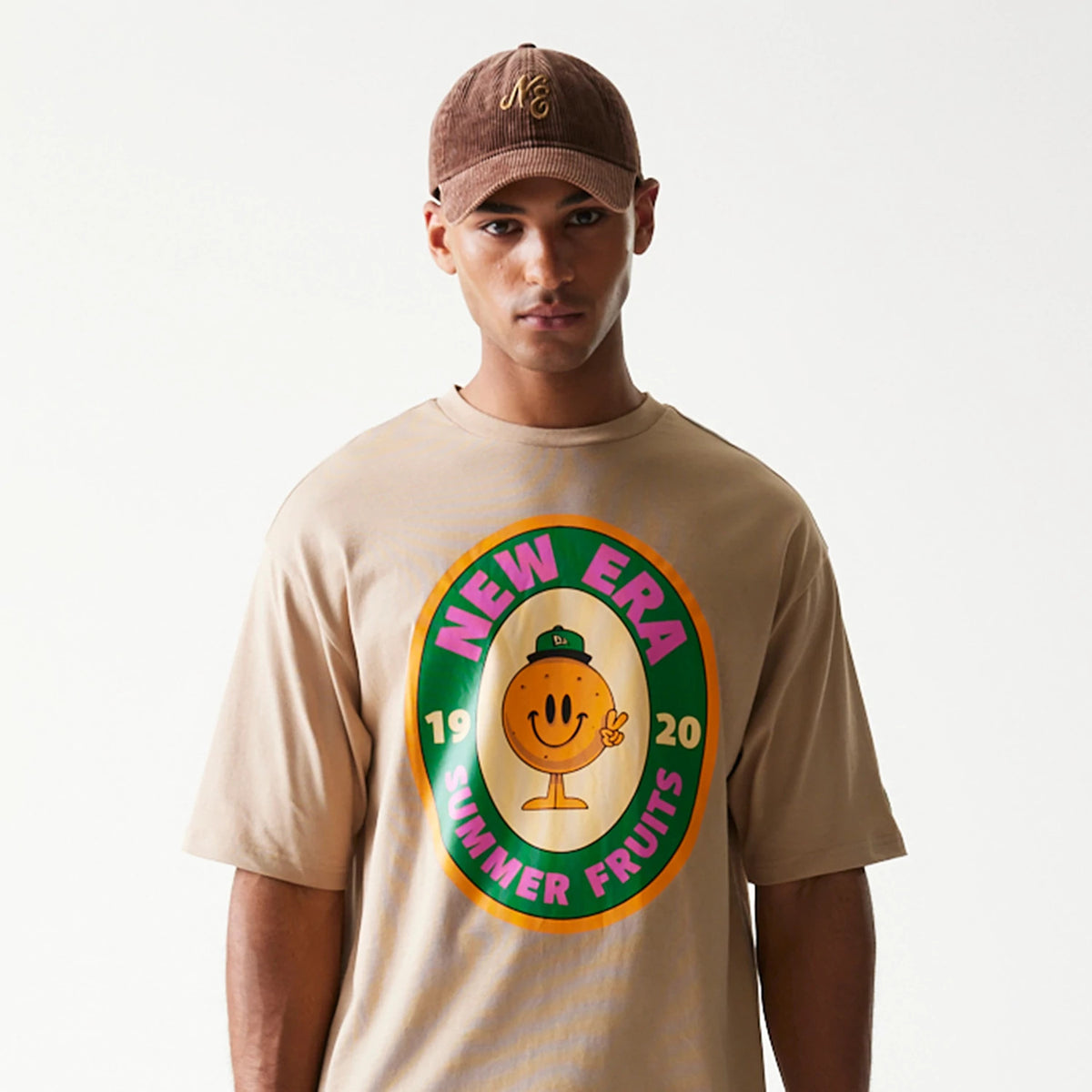 New Era Orange Fruit Graphic Light Beige Oversized T-Shirt, front view on a model wearing a brown cap, featuring a large colourful graphic with New Era Summer Fruits text encircling a smiling fruit character on the chest.