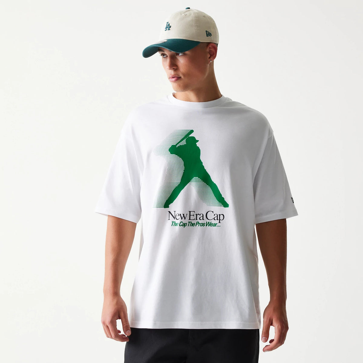 New Era Baseball Player Graphic White Oversized T-Shirt, front view on-model showing a green silhouette of a baseball player mid-swing with the New Era Cap slogan below, short sleeves and a relaxed fit.