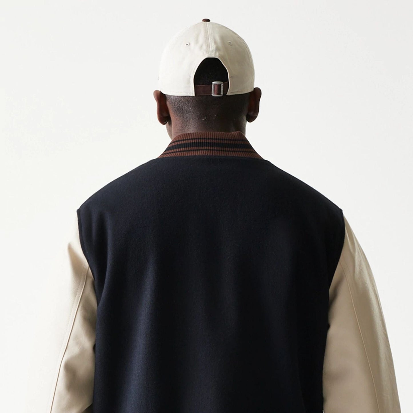 New Era Varsity Black Jacket, back view on-model highlighting the contrasting cream sleeves with a black body, a ribbed collar with brown stripes, and an overall classic varsity silhouette.