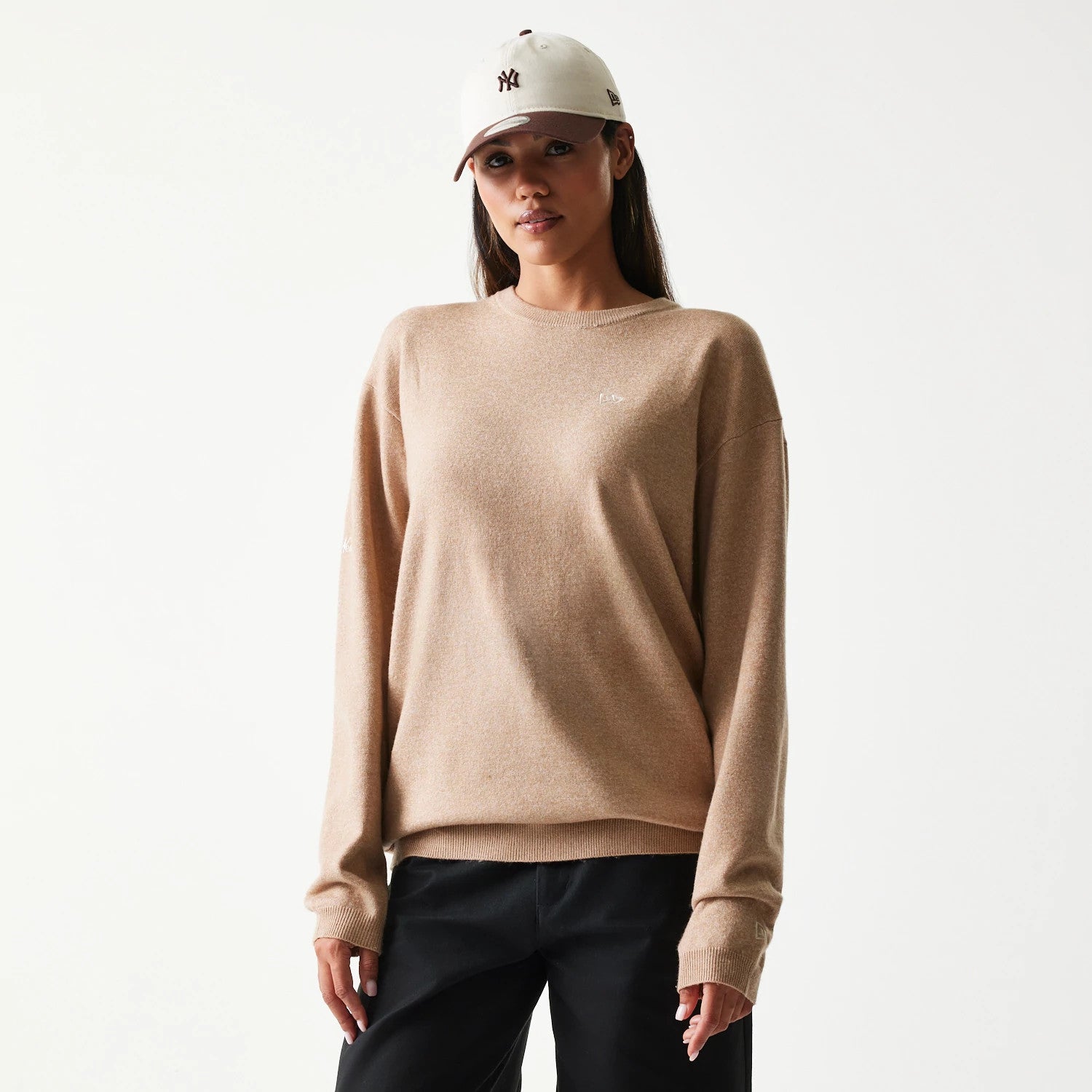 New Era Essential Flag Light Beige Knitted Sweater, front view on a model with a New York Yankees cap, showing the small white embroidered New Era logo on the left chest and right sleeve cuff.