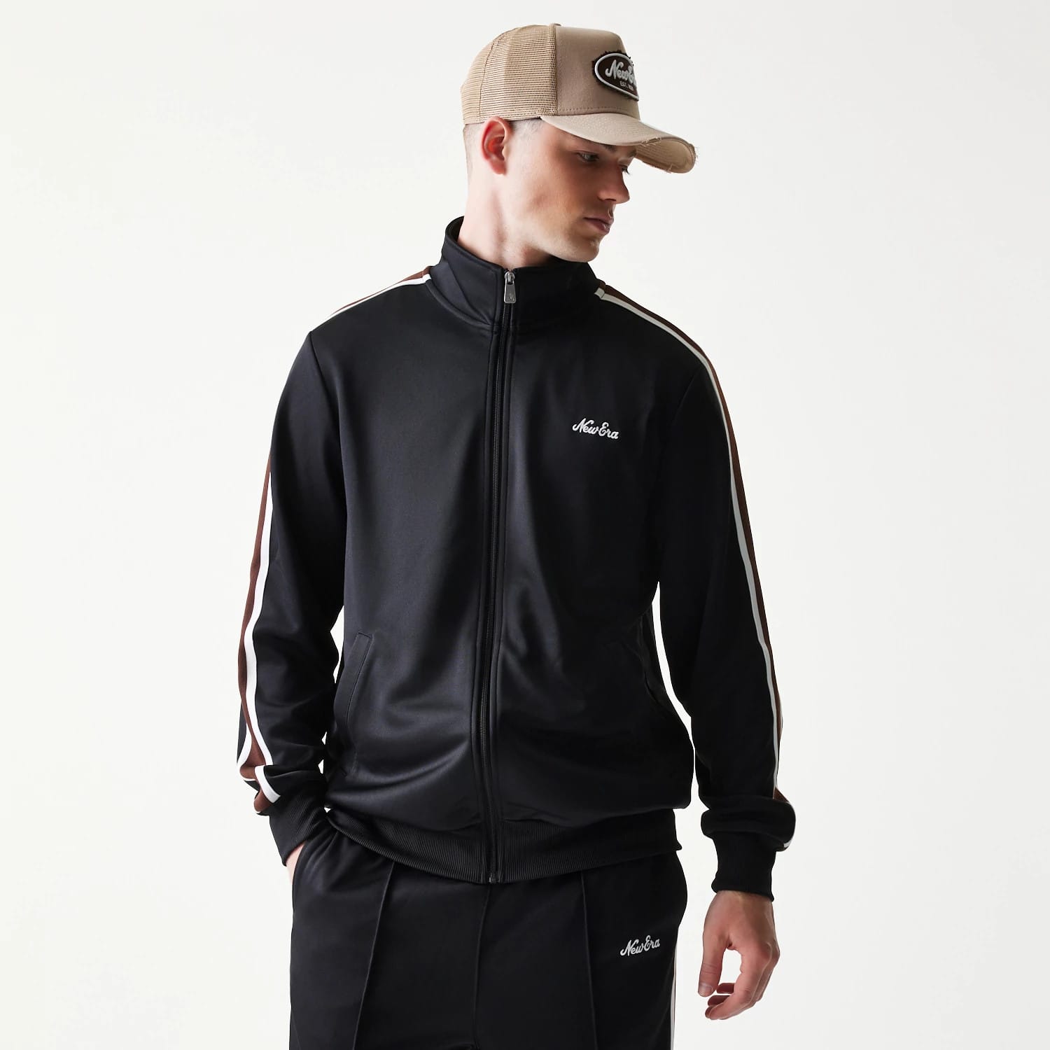 New Era Script Black Track Jacket, front view on a model showing the black jacket with white embroidered script logo on the left chest, zip front, striped sleeves, and matching trousers featuring the same logo on the left leg.