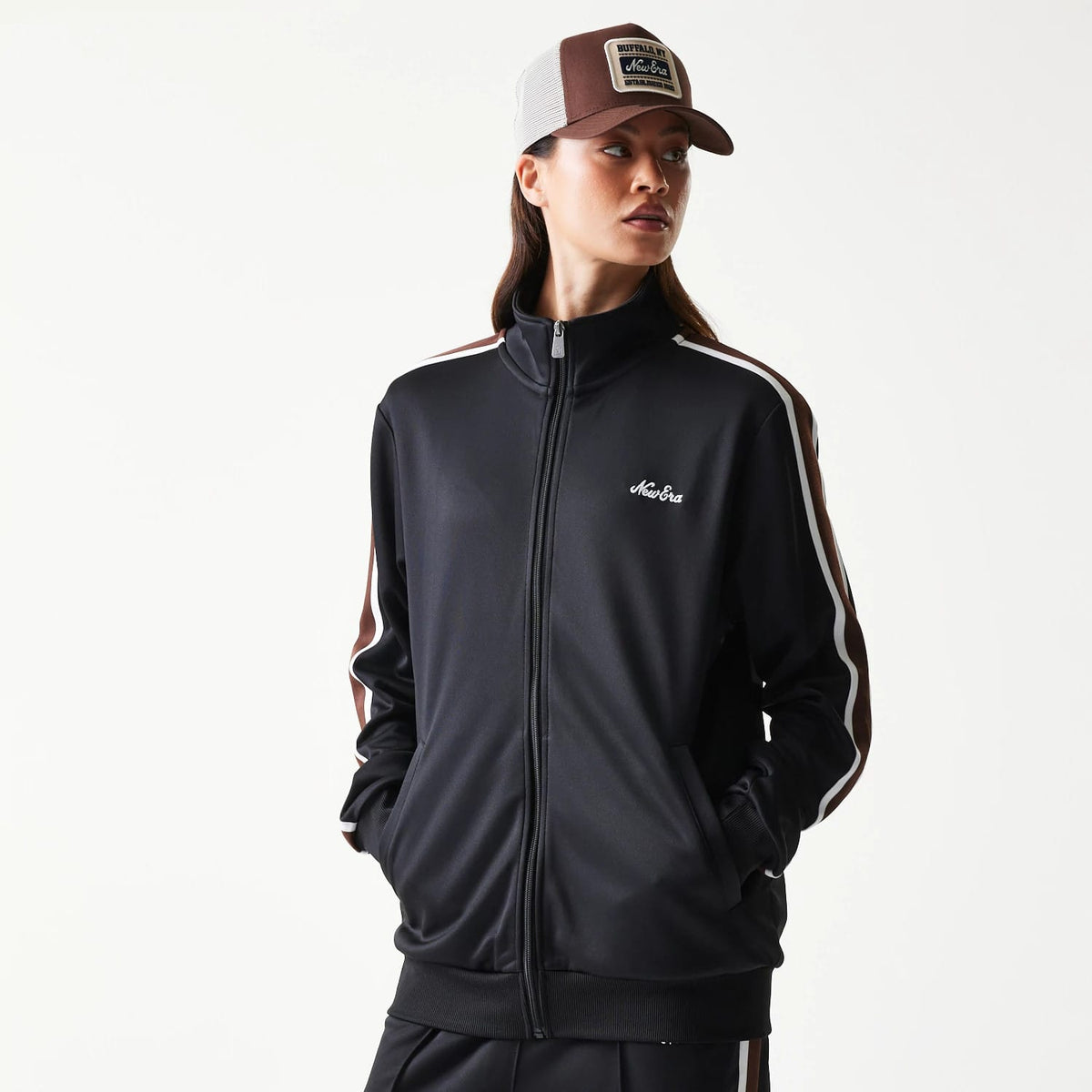 New Era Script Black Track Jacket, front view on a model showing the zipped black jacket with white and brown stripes down the sleeves and an embroidered New Era logo on the left chest, styled with hands in pockets and a matching cap.