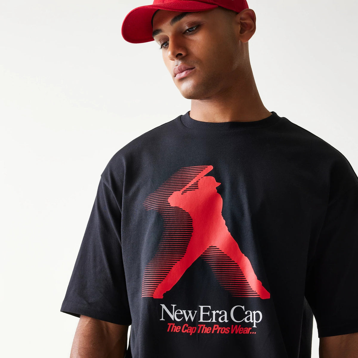 New Era Baseball Player Graphic Black Oversized T-Shirt, front view on-model showing a red silhouette of a baseball player in motion with a bat, New Era Cap branding and the slogan The Cap The Pros Wear printed below the graphic.