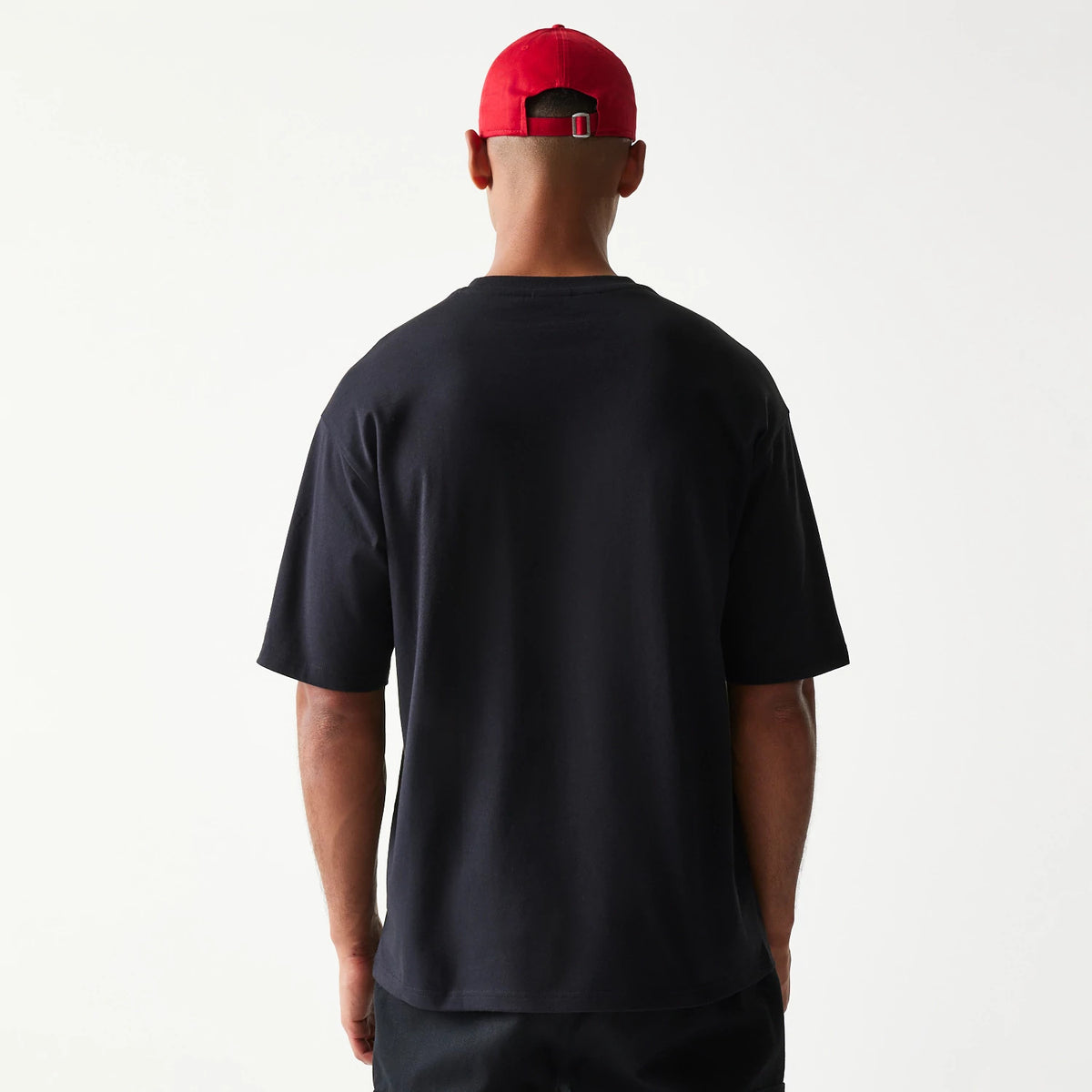 New Era Baseball Player Graphic Black Oversized T-Shirt, back view on a model wearing a red cap, showing the plain black back with a relaxed fit and short sleeves extending just above the elbows.