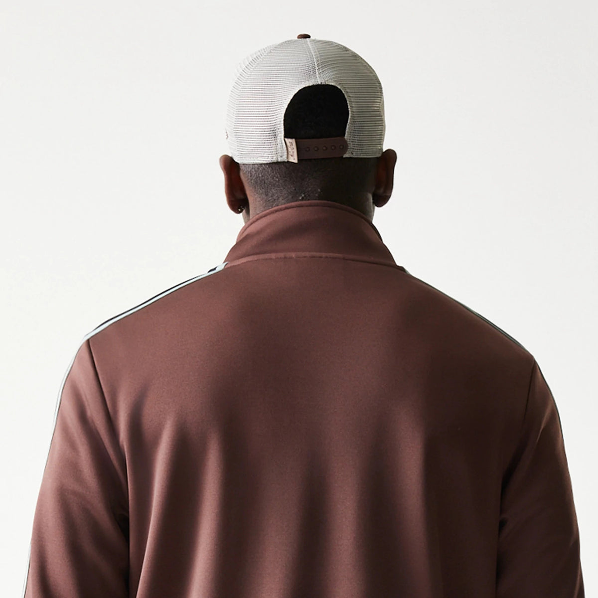 New Era Script Brown Track Jacket, back view on-model displaying simple brown fabric with white shoulder stripes, complemented by a grey cap featuring an adjustable strap at the back.