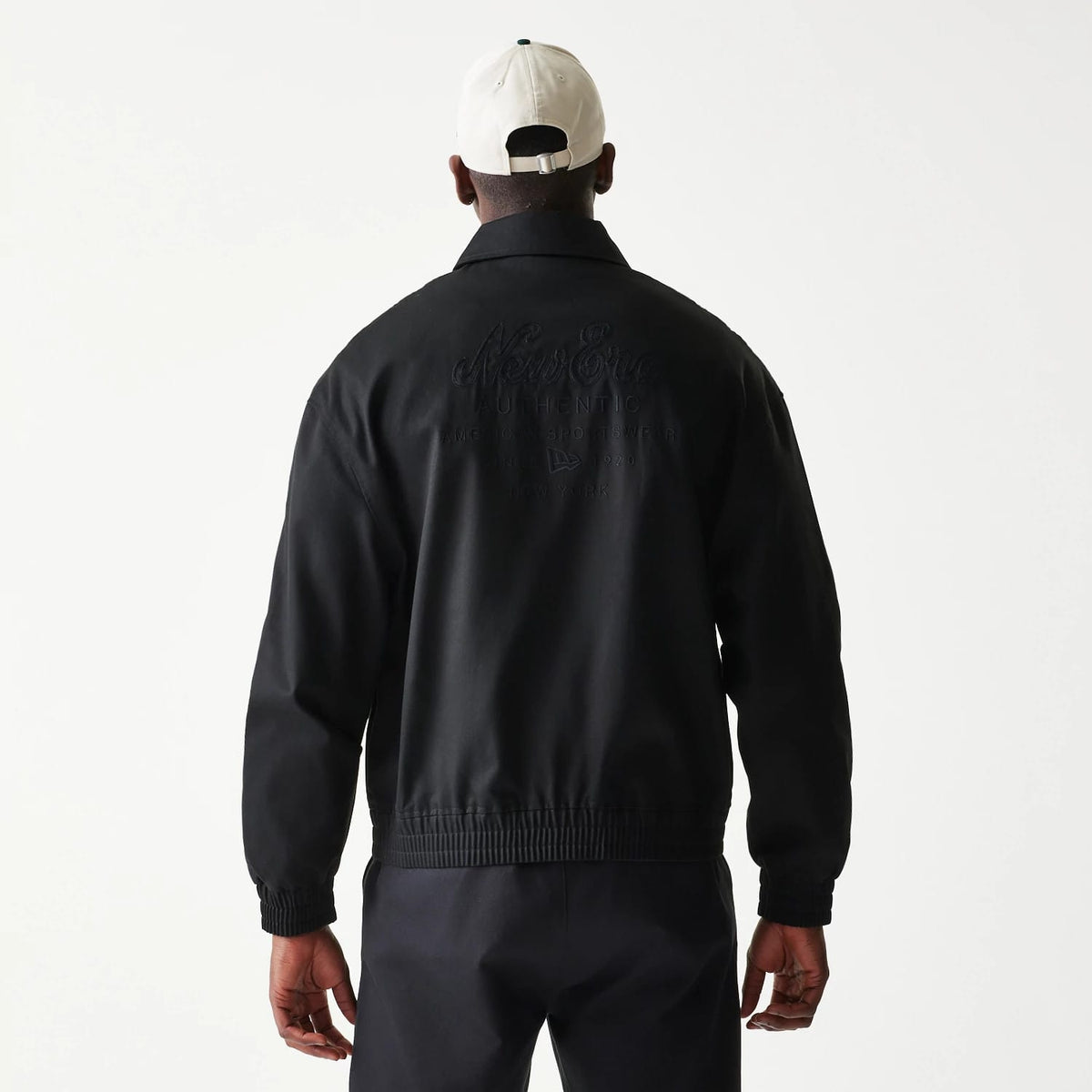 New Era Collared Black Jacket, back view showing the debossed New Era logo and lettering across the upper back, worn by a model styled with dark trousers and a complementary light-coloured cap.