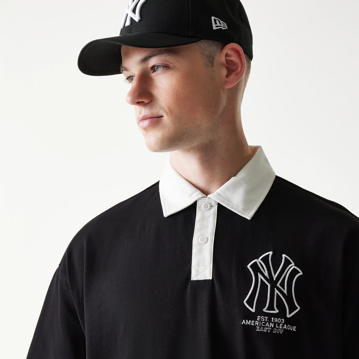 New Era New York Yankees MLB Black Oversized Polo T-Shirt, three-quarter view on a model styled with a black cap, featuring a prominent embroidered white NY logo on the left chest and a contrasting white collar with button placket.