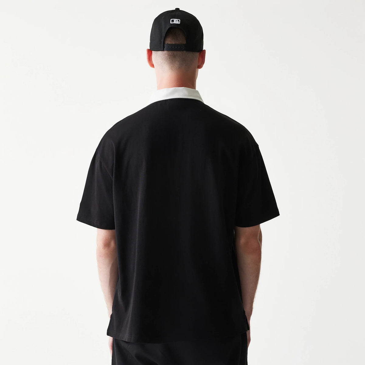 New Era New York Yankees MLB Black Oversized Polo T-Shirt, back view on-model showing a white contrast collar with a loose fit, paired with a black cap featuring an embroidered MLB logo on the rear of the cap.