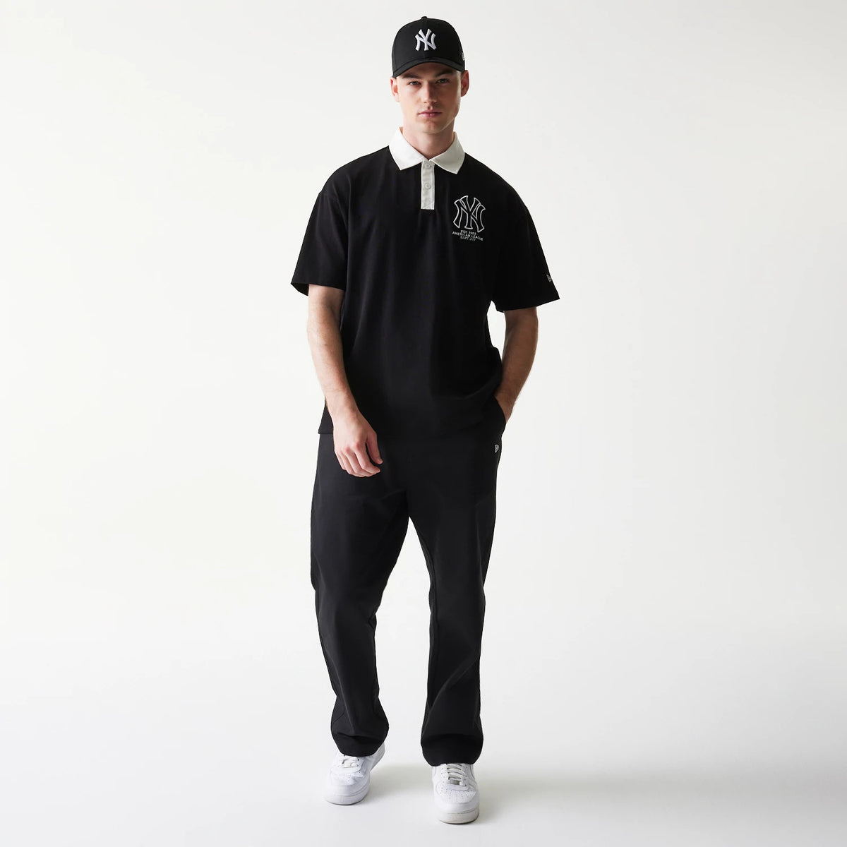 New Era New York Yankees MLB Black Oversized Polo T-Shirt, front view on a model styled with matching black trousers and a cap, featuring a large embroidered NY logo on the chest and a contrasting white collar.
