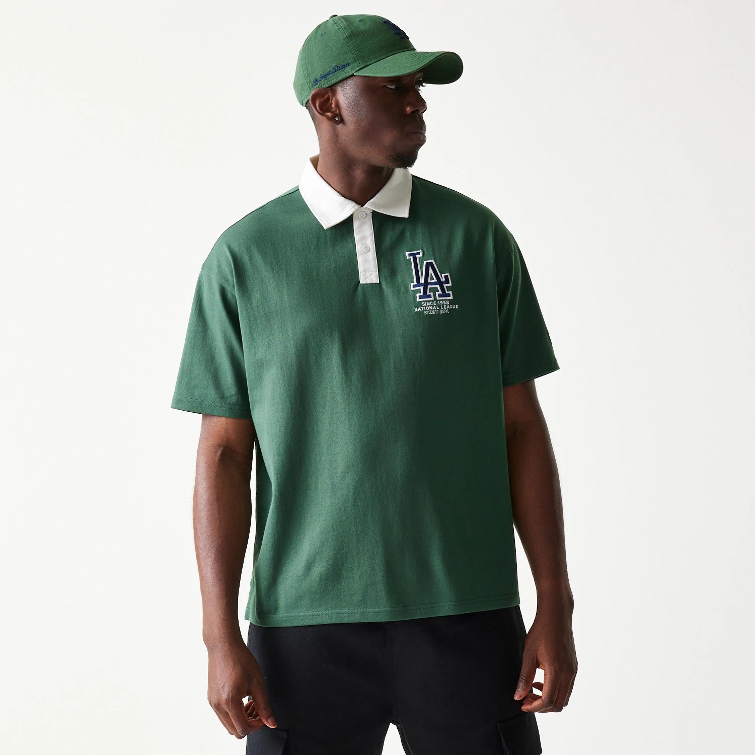 New Era LA Dodgers MLB Dark Green Oversized Polo T-Shirt, front view on model showing the embroidered LA logo in white and navy on the left chest, contrasted by a white collar, styled with a matching dark green cap.