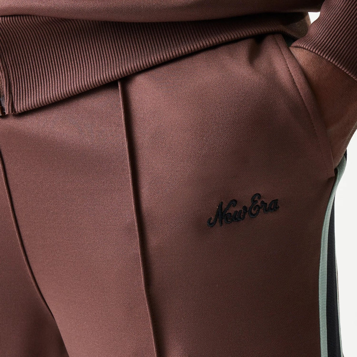 New Era Script Brown Straight Leg Joggers, close-up showing the black embroidered New Era script logo on the brown fabric above the front pocket, with contrasting side stripe running down the leg.