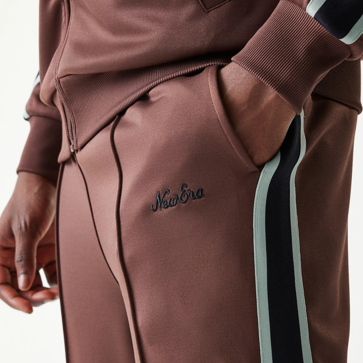 New Era Script Brown Straight Leg Joggers, close-up view showing black embroidered New Era script on the thigh, with detailed striped trim down the side and ribbed cuffs, highlighting the pocket and zip on a matching brown jacket.